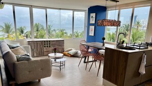 A modern living room with a sofa, a dining table, and chairs, a kitchen area with a sink and stove, and large windows offering a view of palm trees and a cityscape. - U-go Faro Tequendema (Cartagena)