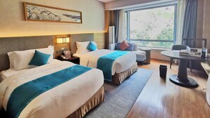A hotel room with two beds, a desk, a chair, a small table, and a large window with a view of trees. - Crowne Plaza Hangzhou Thousand Island Lake (Hangzhou)
