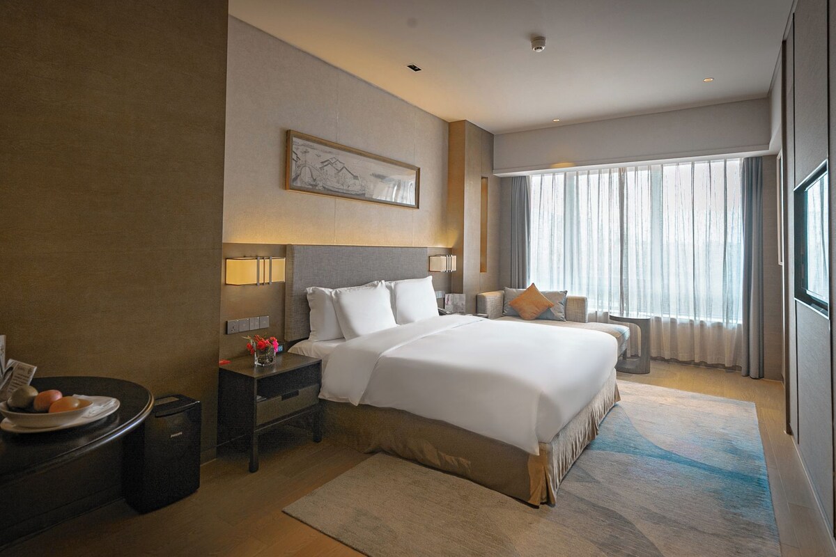 Quarto de hotel com uma cama grande, mesas de cabeceira, luminária, um quadro na parede e uma janela com cortinas.