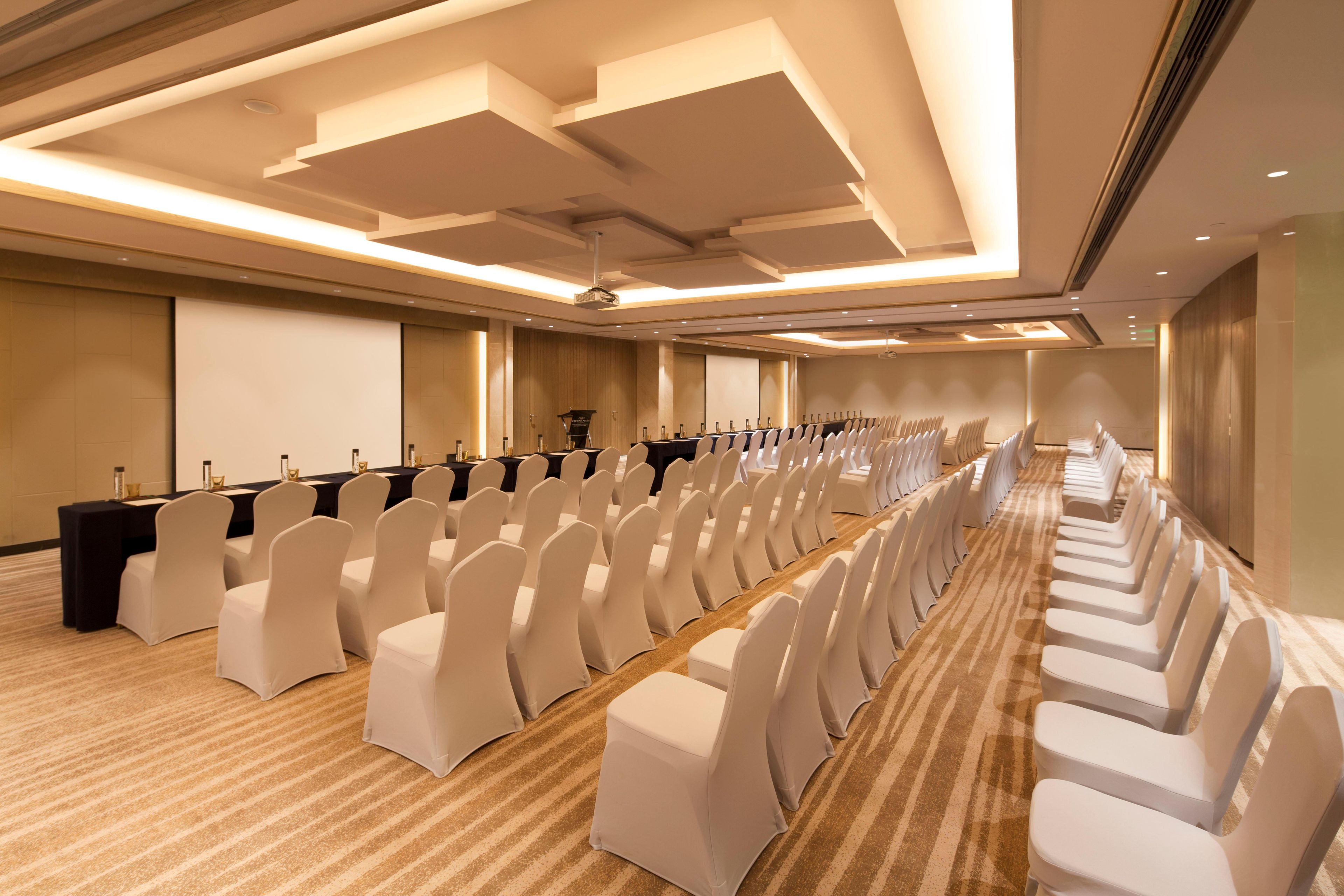 Ein Konferenzraum mit Reihen weißer Stühle, einem langen Tisch und einer Projektionsleinwand.