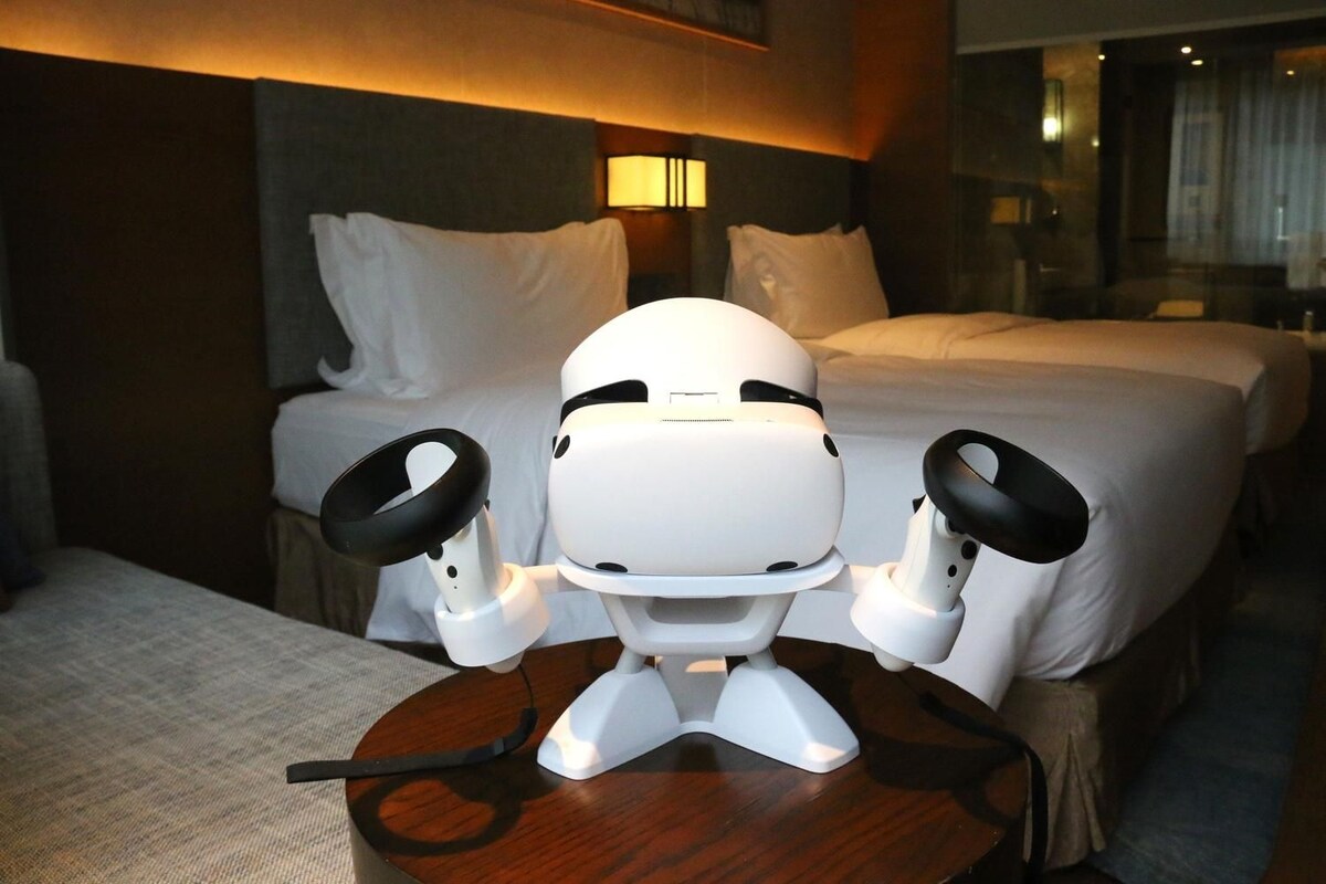 Um robô com duas peças circulares pretas sobre uma mesa num quarto de hotel com duas camas e uma mesinha de cabeceira.