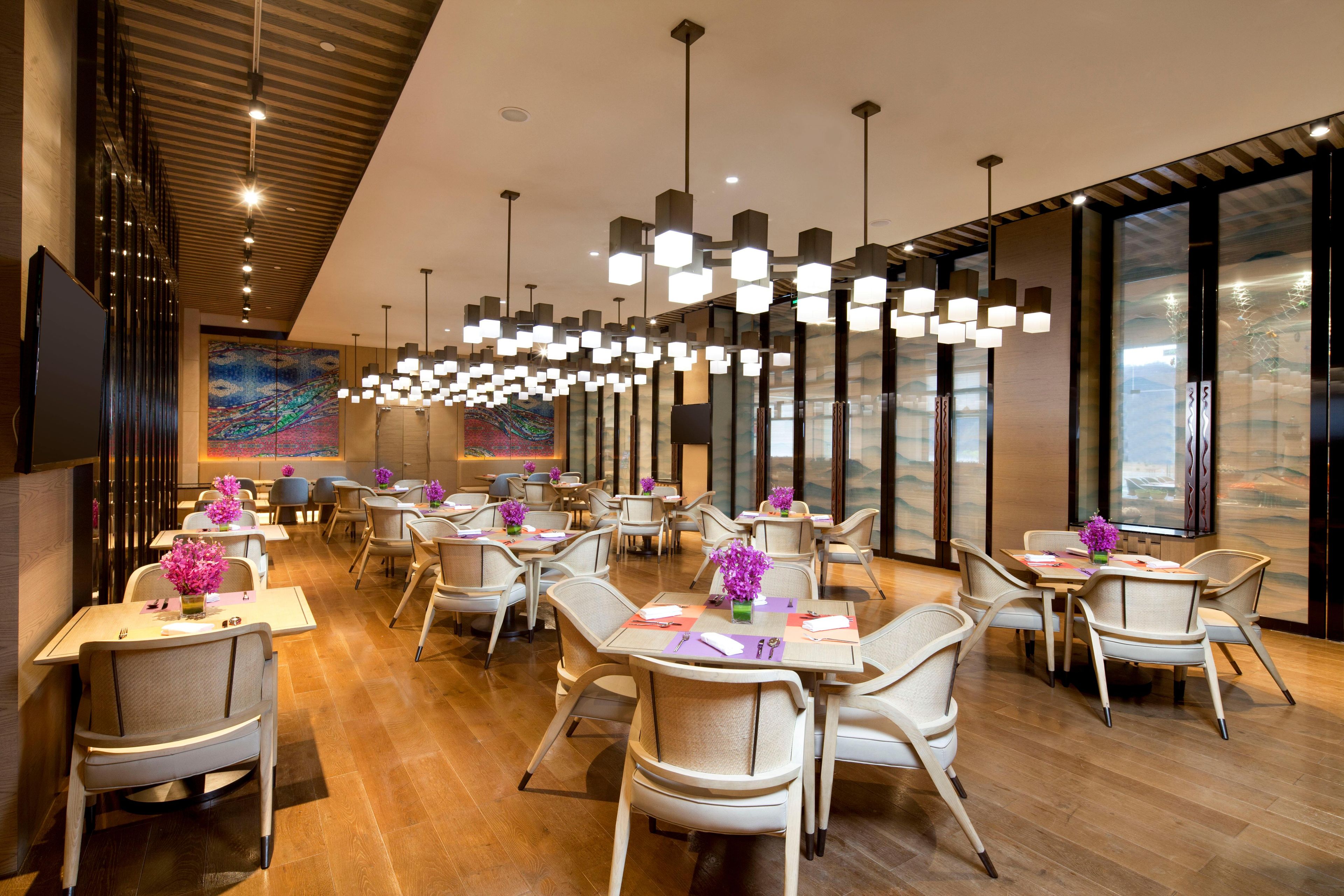 A modern restaurant with multiple tables and chairs, pendant lighting, and large windows.