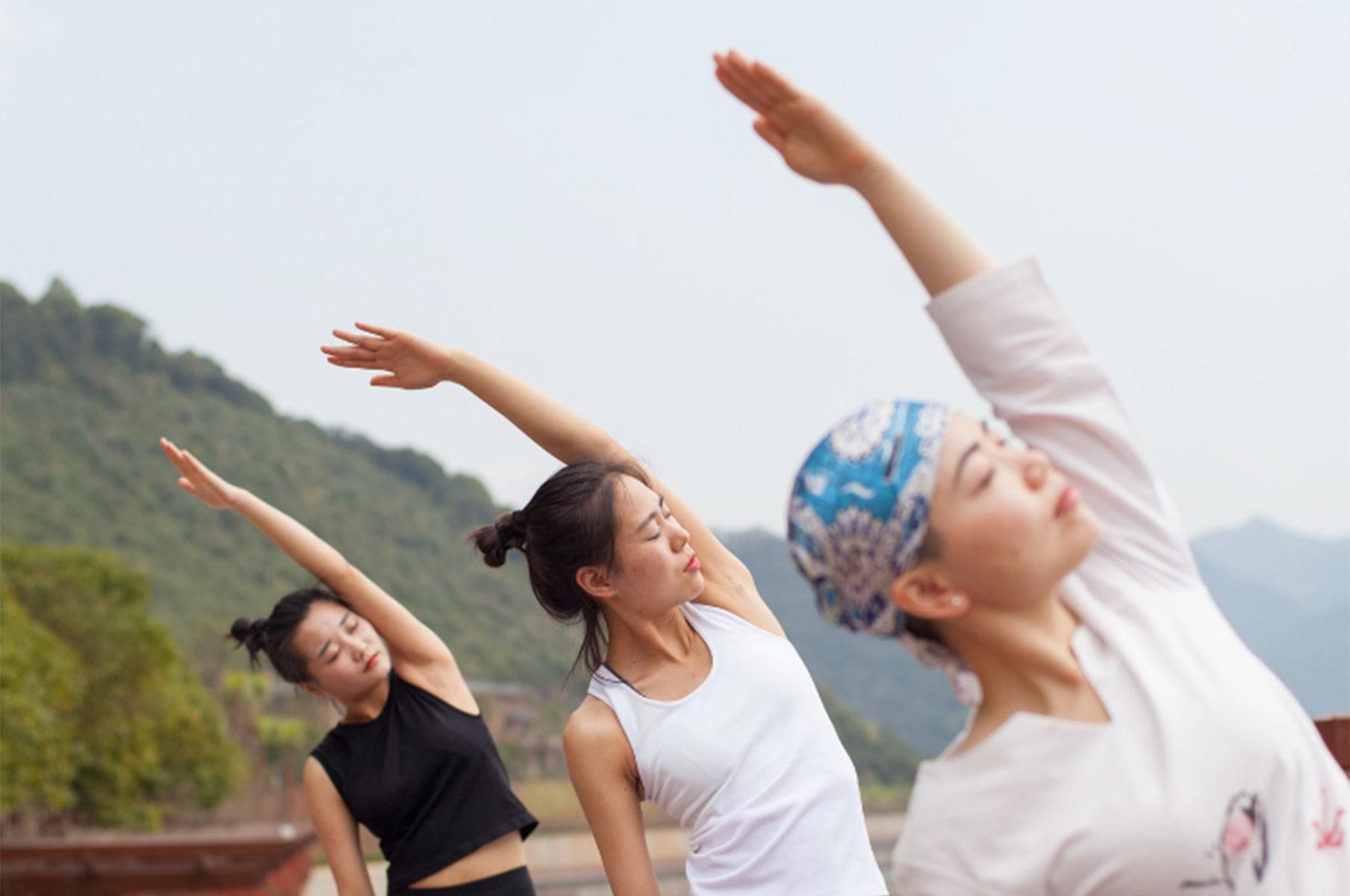 Tres personas realizan una postura de yoga al aire libre, con montañas al fondo.