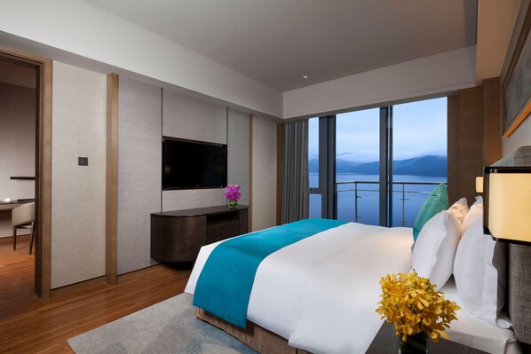 A modern hotel room with a large bed, a flat-screen TV, a desk, and a view of the sea.