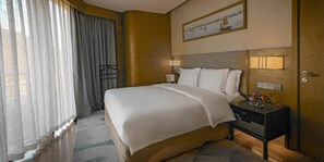 A hotel room with a large bed, a nightstand with lamps, a painting on the wall, and a view of the outdoors through sheer curtains.