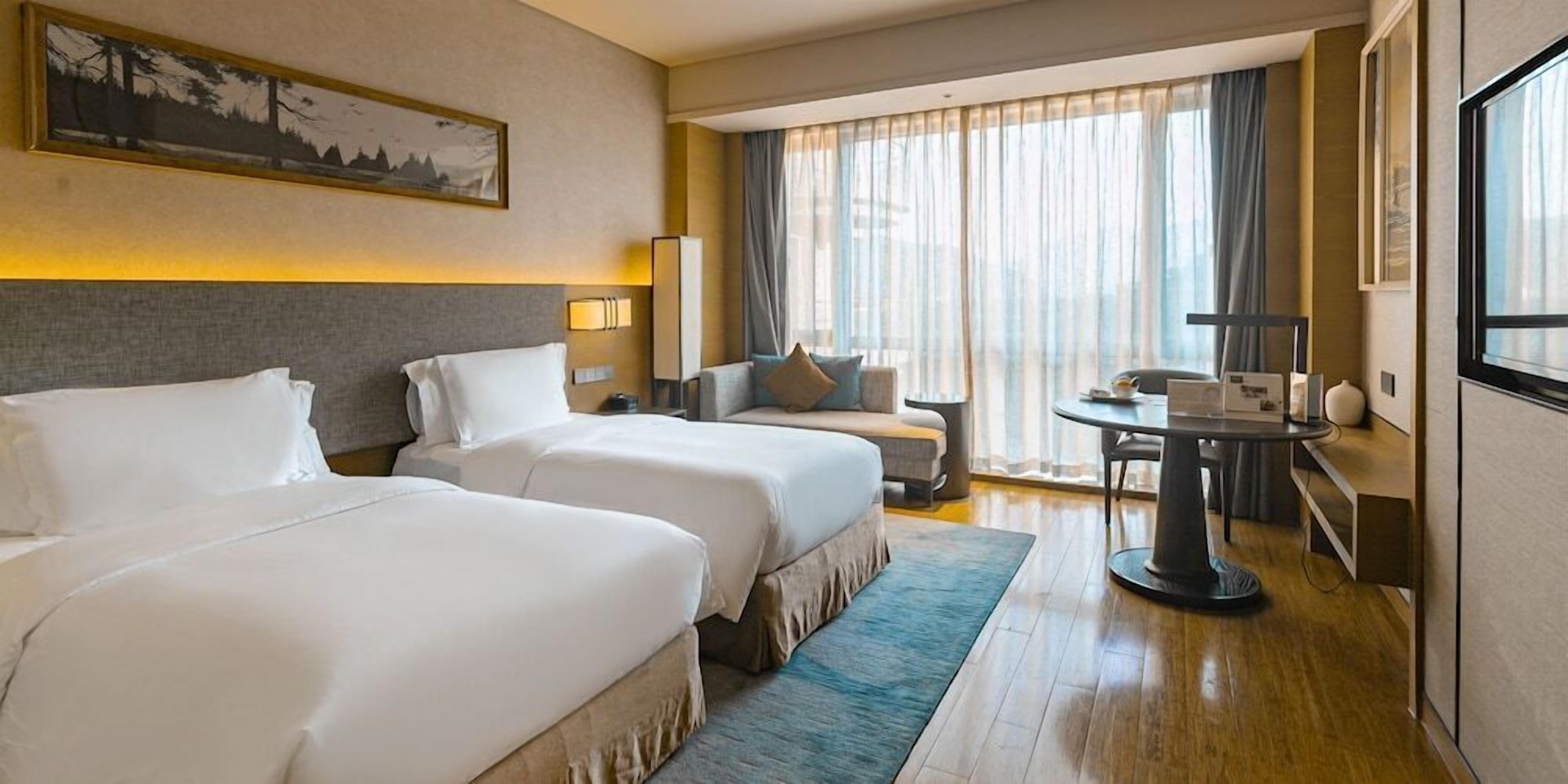 A hotel room with two beds, a small table, a sofa, and a large window with curtains.