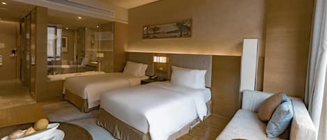 A hotel room with two beds, a sofa, a dining table with a bowl of fruit, a mirror, and a painting on the wall.