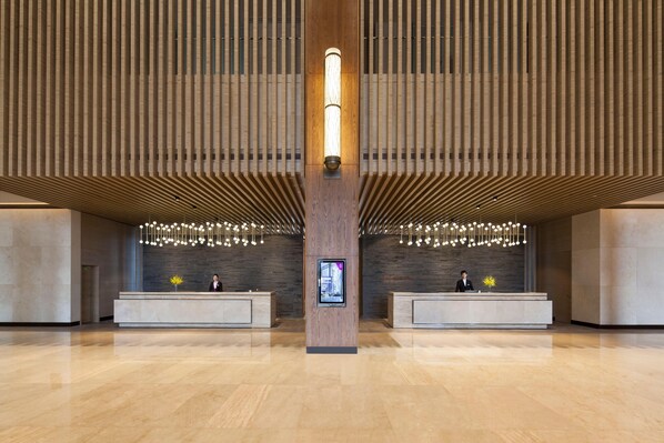 A spacious lobby with a central reception desk, wooden paneling on the walls, and a modern chandelier.