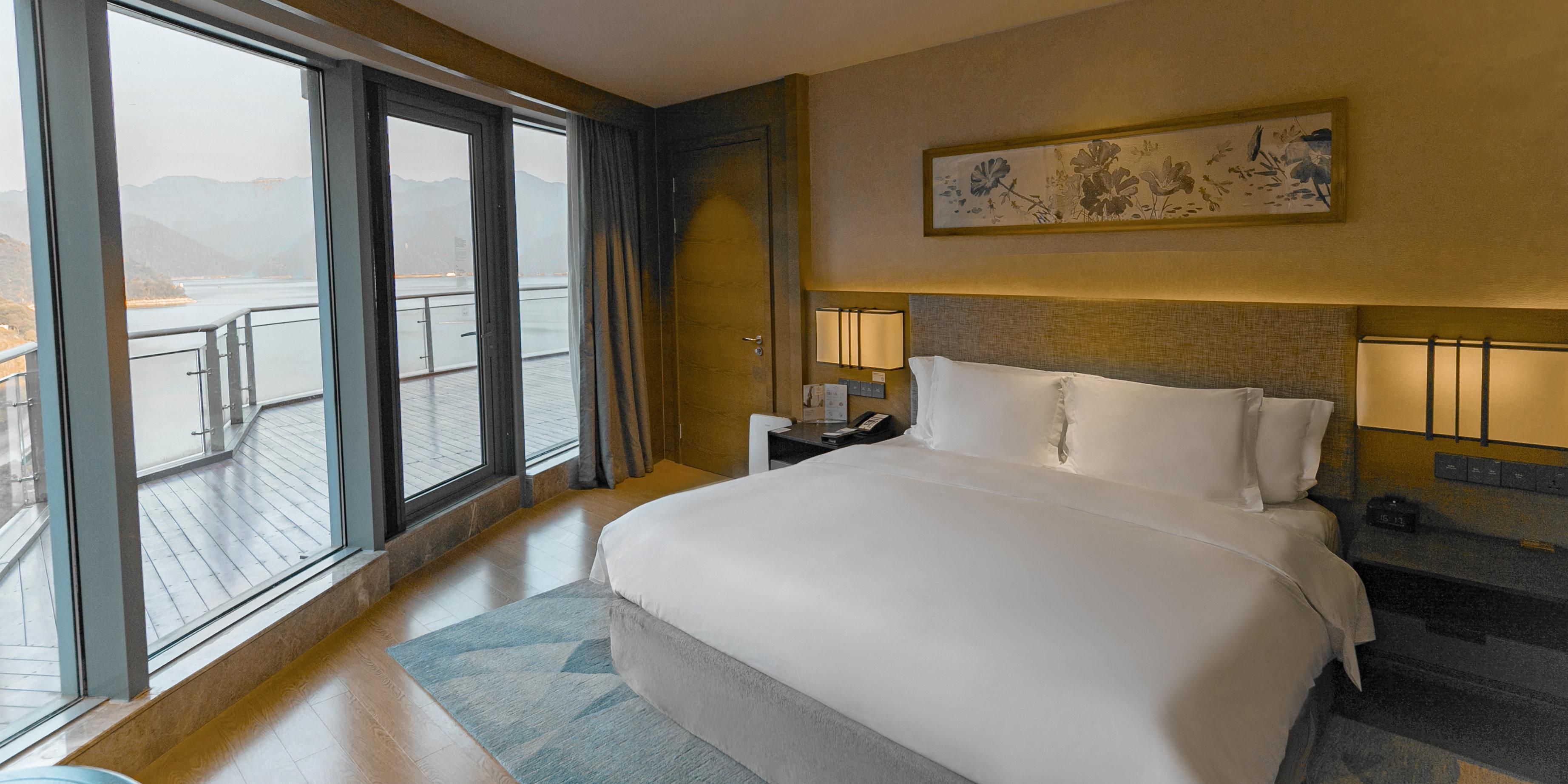 A hotel room with a large bed, a nightstand, a lamp, and a view of a body of water through a sliding door.