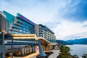 Un hotel moderno con una entrada curva destacada, una fuente de agua grande y vistas a un lago y montañas.