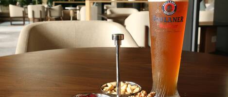 Un vaso alto de cerveza con un logotipo, un recipiente metálico para mantequilla y dos cuencos metálicos con frutos secos y frutas deshidratadas sobre una mesa de madera.