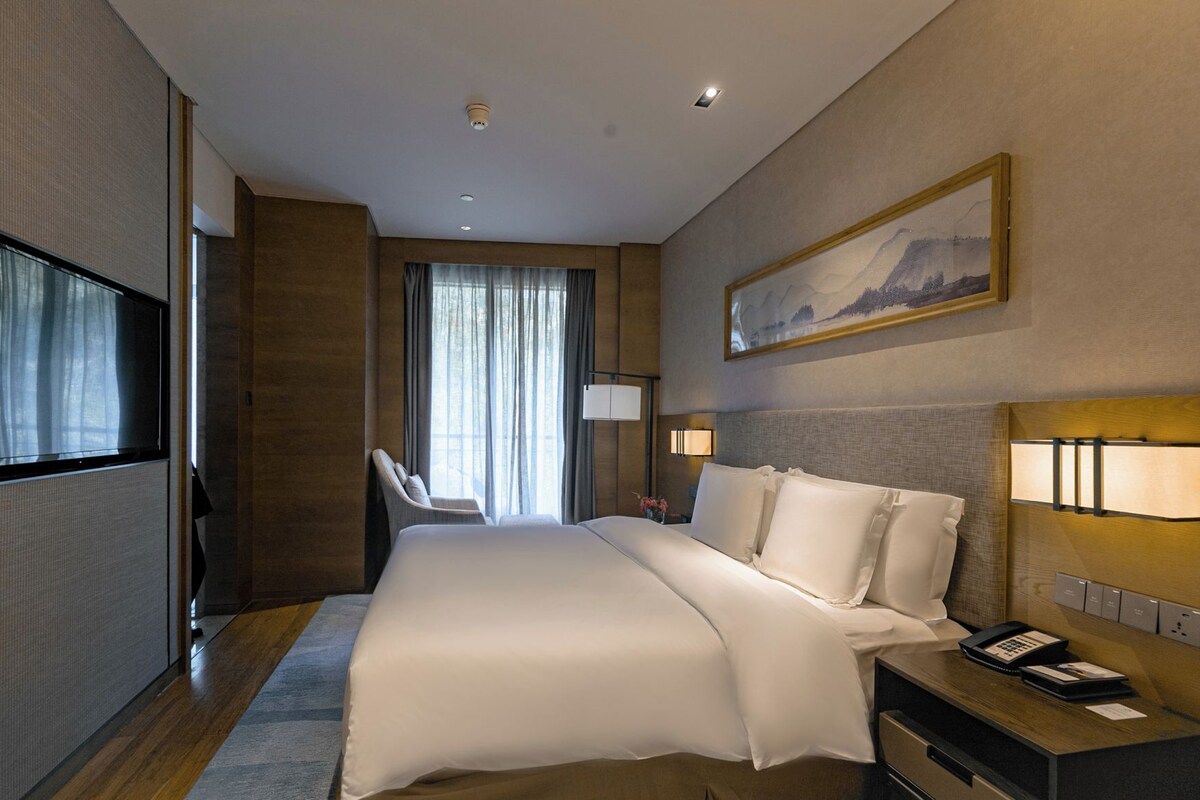Quarto de hotel com uma cama grande, uma escrivaninha com telefone, uma cadeira, uma televisão e um quadro na parede.