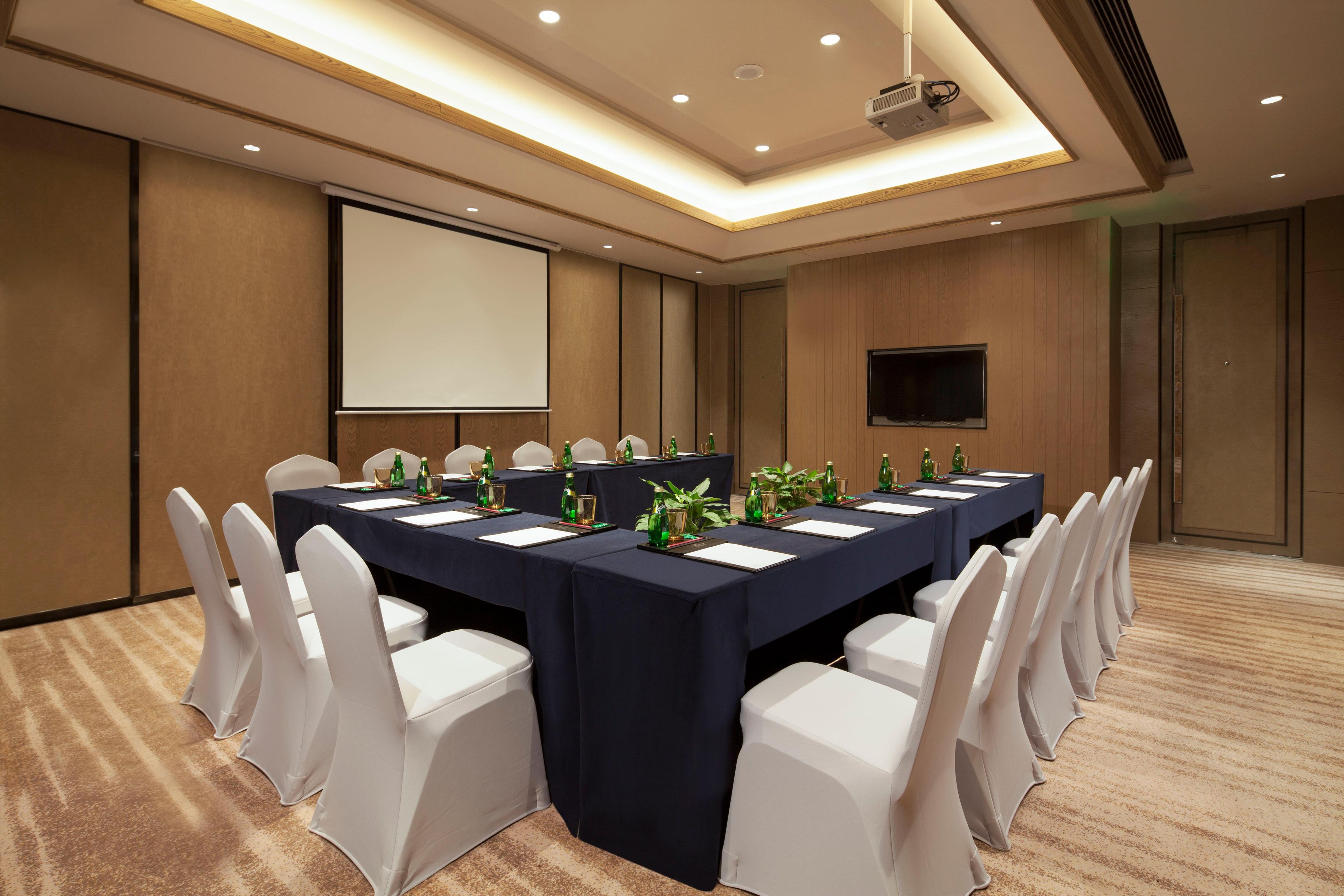 Un salón de conferencias con una larga mesa, sillas blancas, una pantalla de proyector y un televisor de pantalla plana.