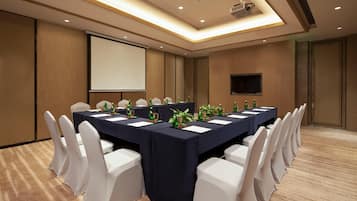 Un salón de conferencias con una larga mesa, sillas blancas, una pantalla de proyector y un televisor de pantalla plana.