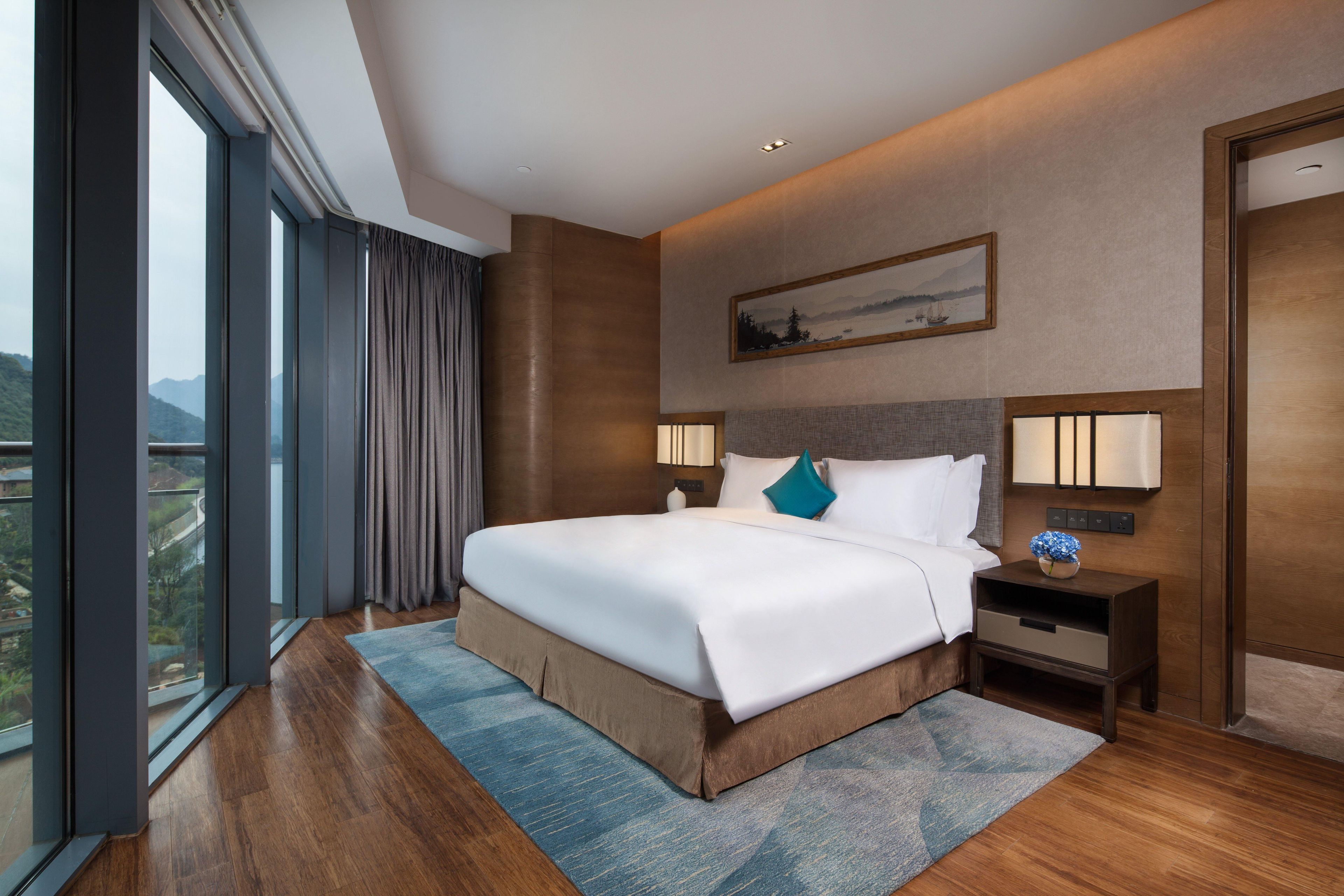 Una habitación de hotel con una cama grande, una mesita de noche, una lámpara de pared y un ventanal con cortinas.