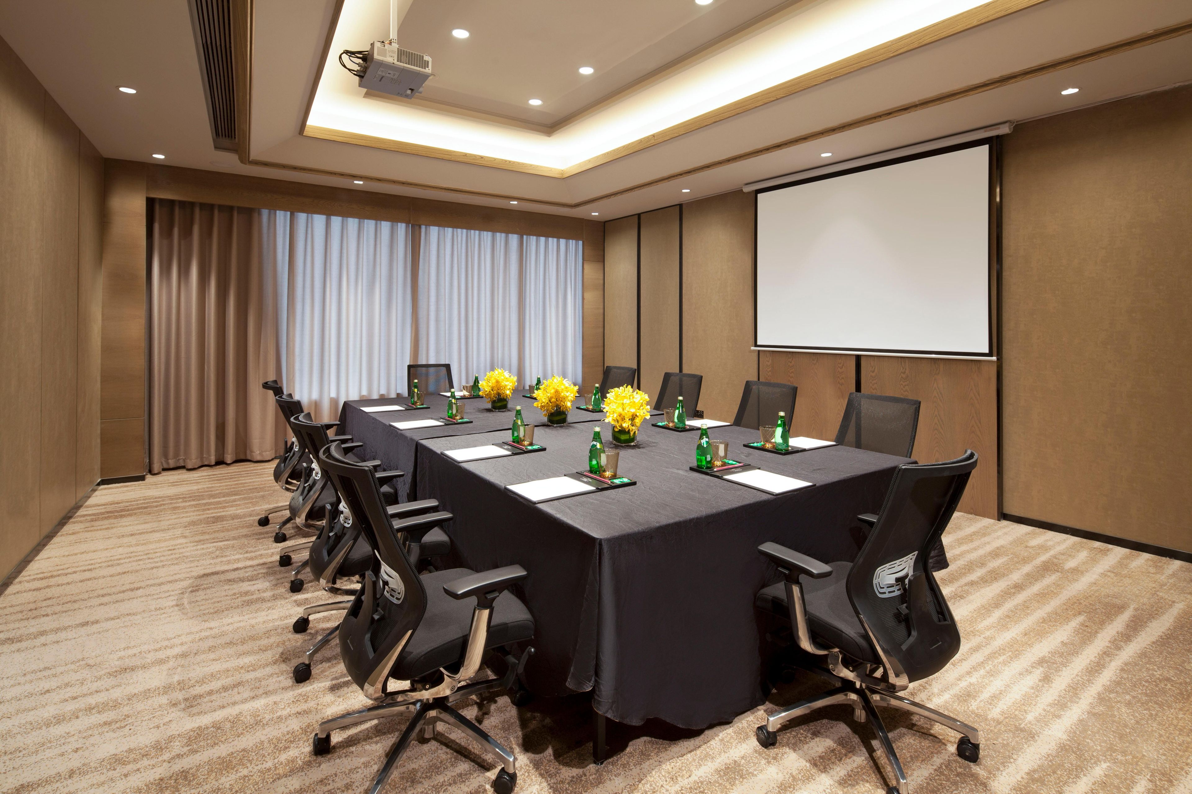 A conference room with a long table, chairs, a projector screen, and floral centerpieces.