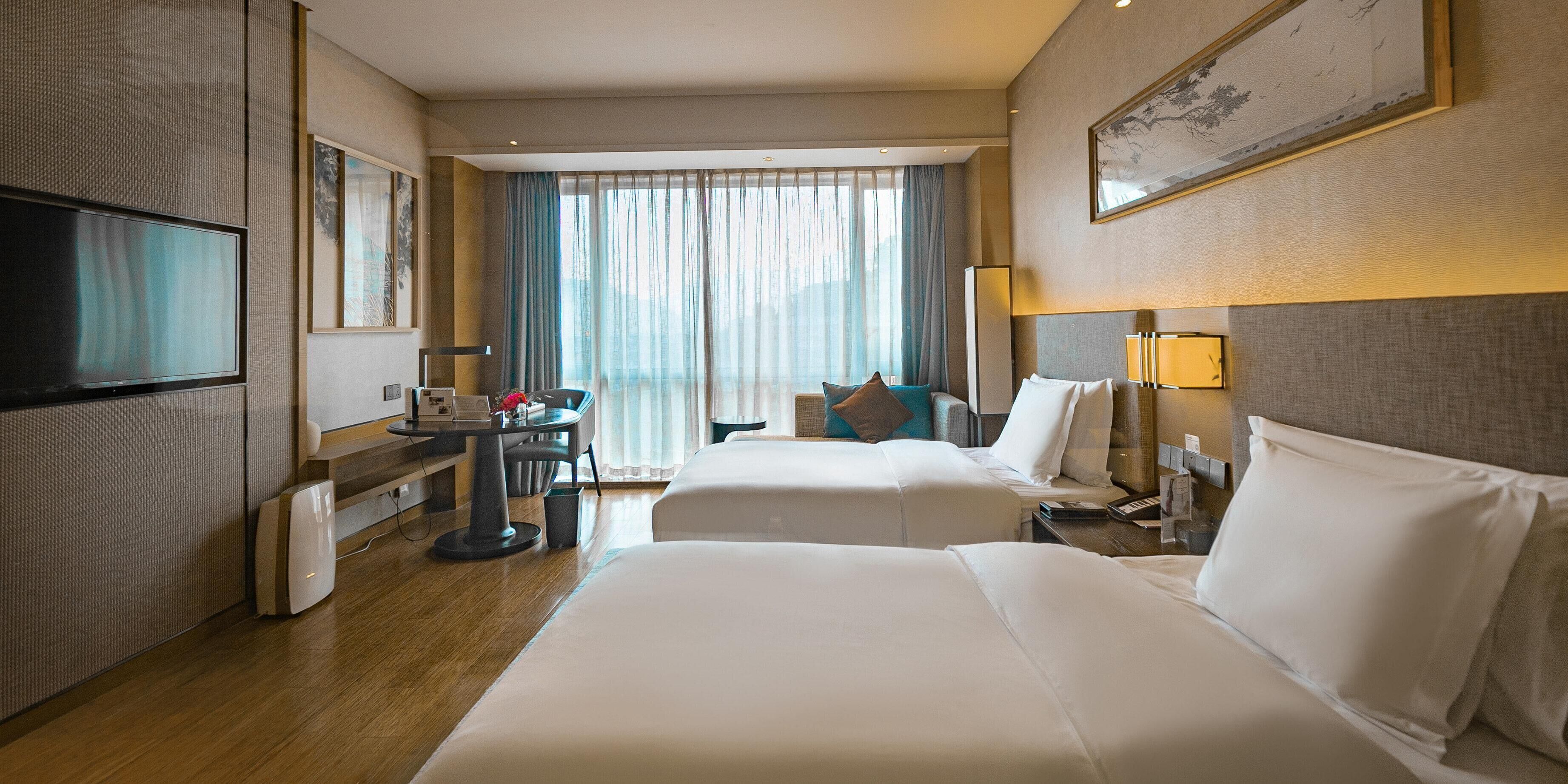 A hotel room with two beds, a desk, a TV, and a window with curtains.
