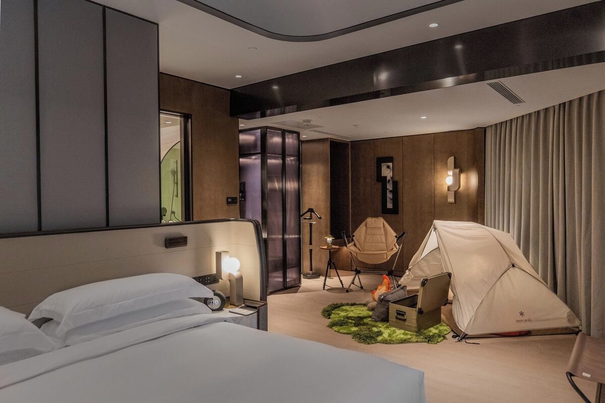 Una habitación de hotel moderna con una cama, una silla, una tienda de campaña y una pequeña mesa con una lámpara.
