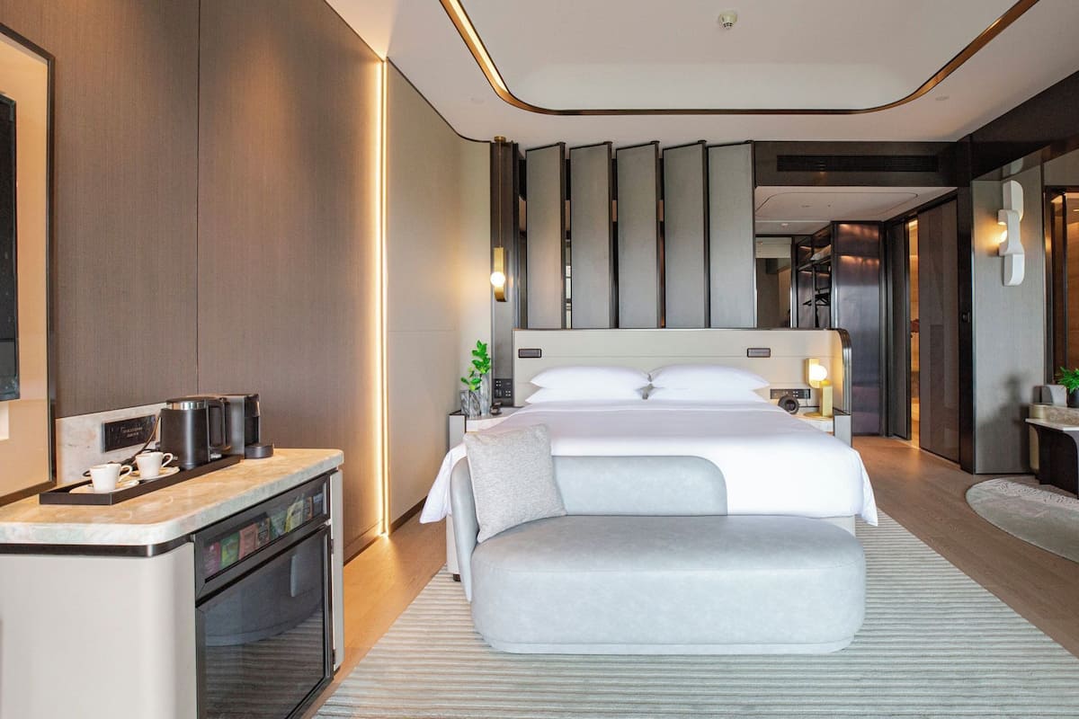 A modern hotel room with a bed, bedside table, and a small kitchenette.
