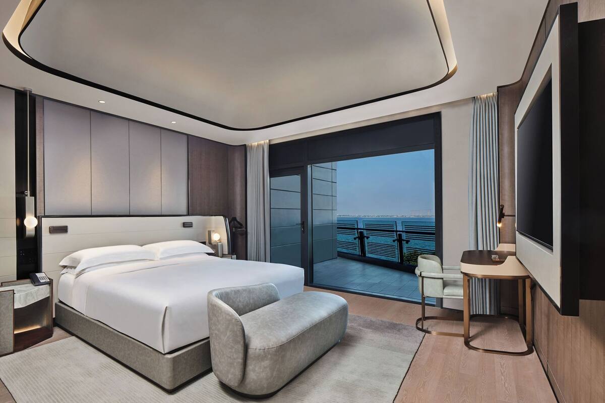 A modern bedroom with a large bed, bedside tables, a seating area, and a view of the sea.