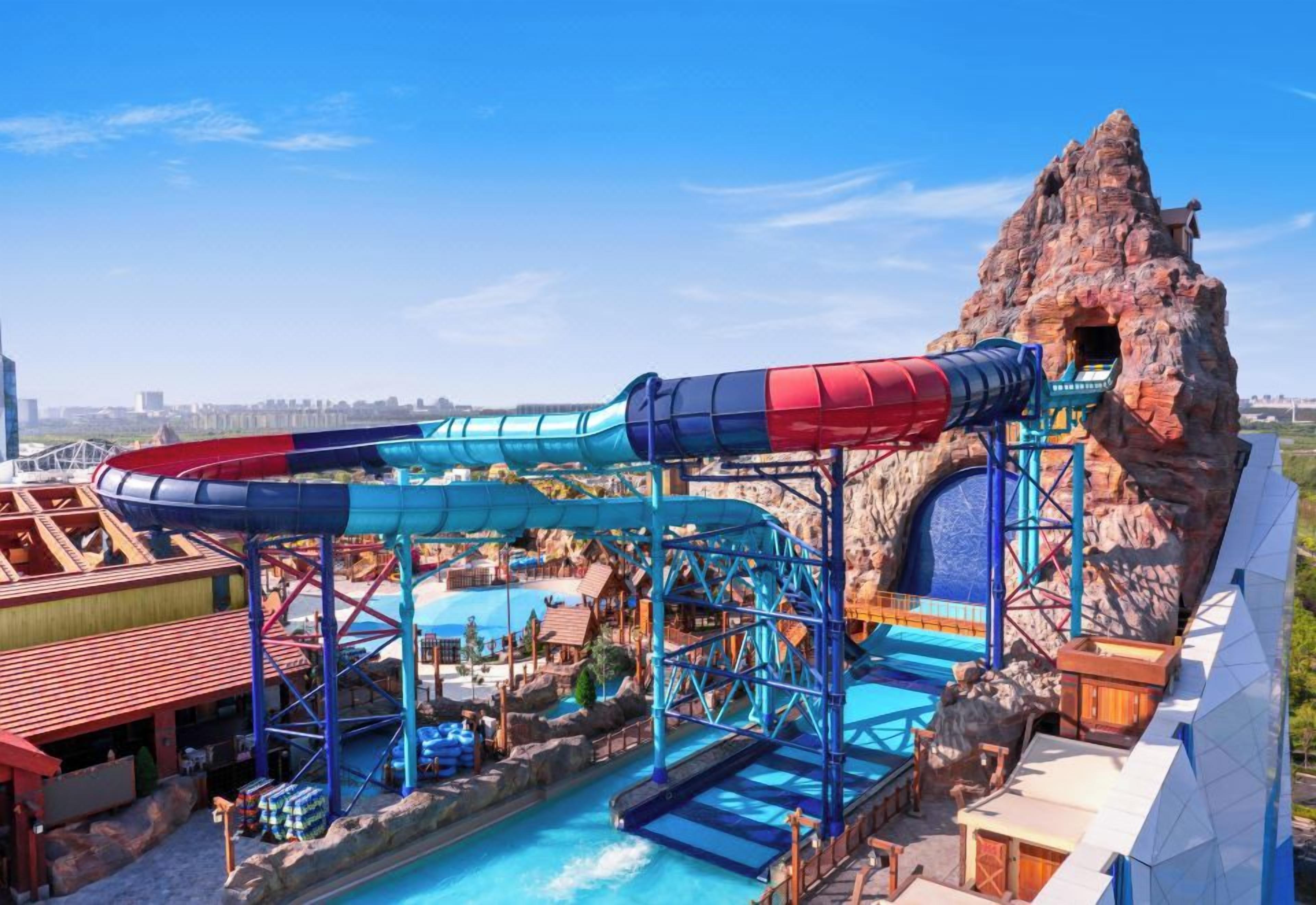 Un parque acuático con un tobogán gigante, una piscina y montañas rocosas al fondo.