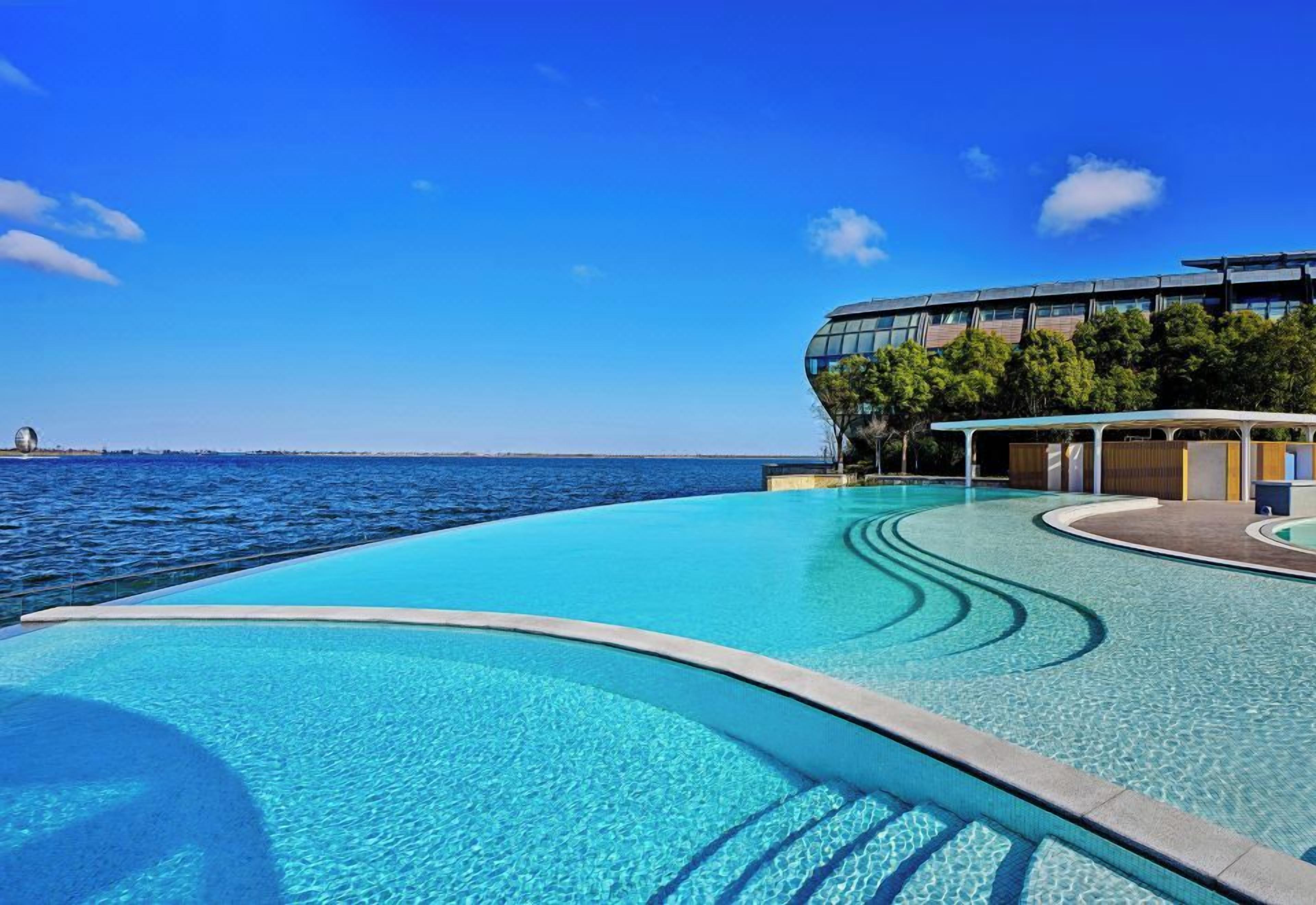 A swimming pool with a curved edge, a modern building with large windows, and a view of the sea.