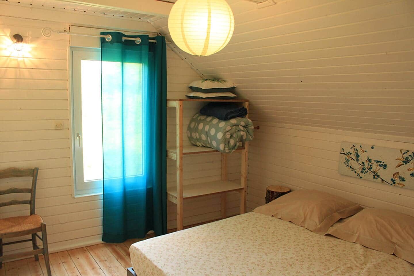 A room with a bed, a chair, a shelf with pillows, a hanging light, and a window with curtains.