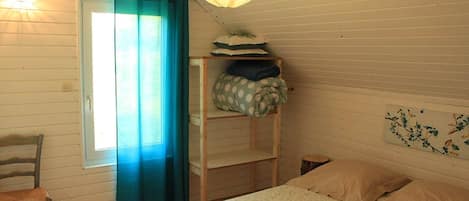 Una habitación con una cama, una silla, un estante con almohadas, una lámpara colgante y una ventana con cortinas.