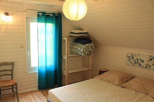 A room with a bed, a chair, a shelf with pillows, a hanging light, and a window with curtains.