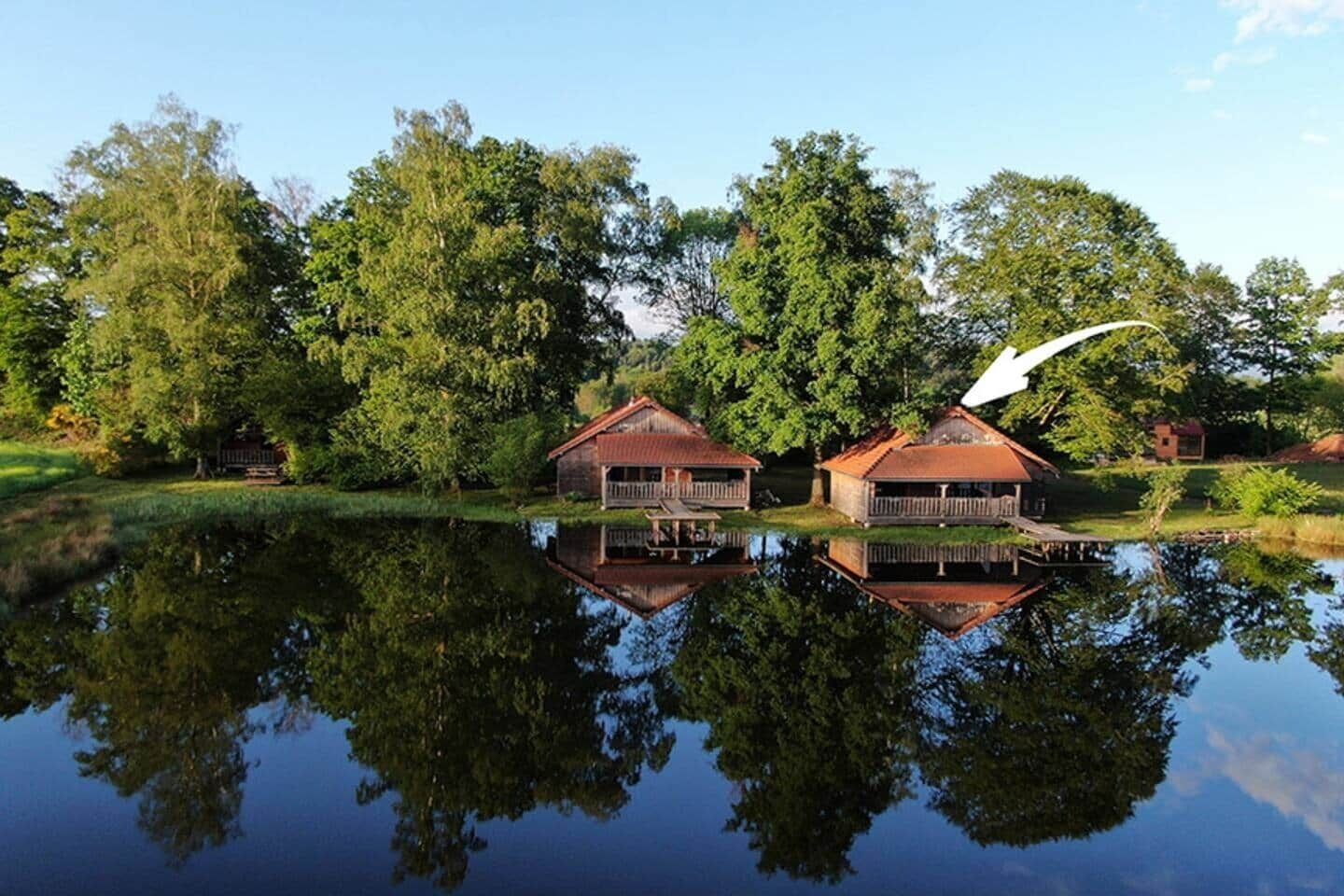 Un cadre paisible au bord d’un lac, avec deux chalets en bois, chacun doté d’un balcon et d’une petite terrasse, le tout entouré d’une végétation luxuriante et d’arbres.
