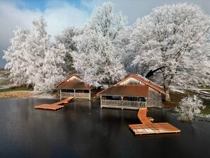 Twee houten hutten met aanleunsteigers aan een rustig meer, omringd door bevroren bomen.