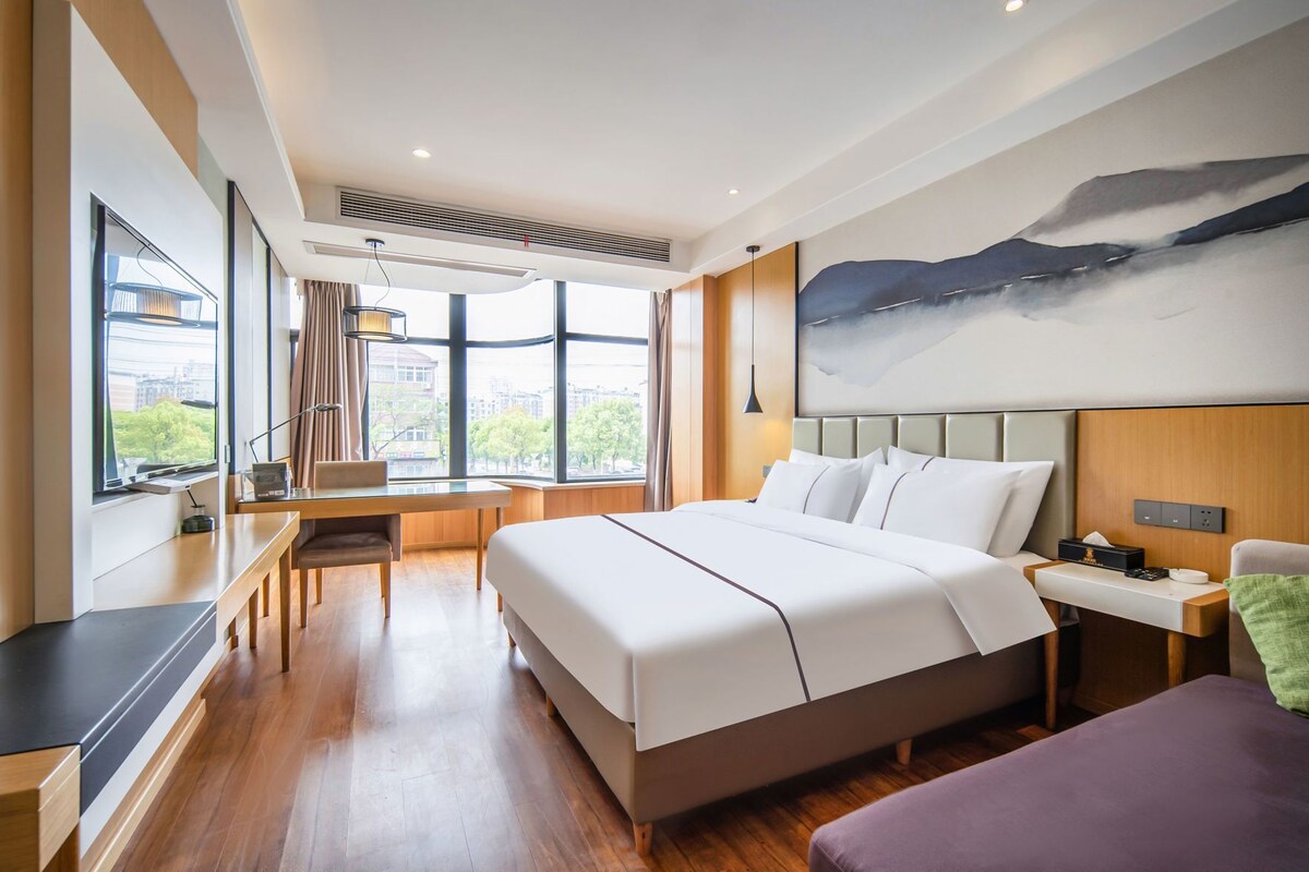A modern hotel room with a large bed, a desk, and a view of the city through large windows.