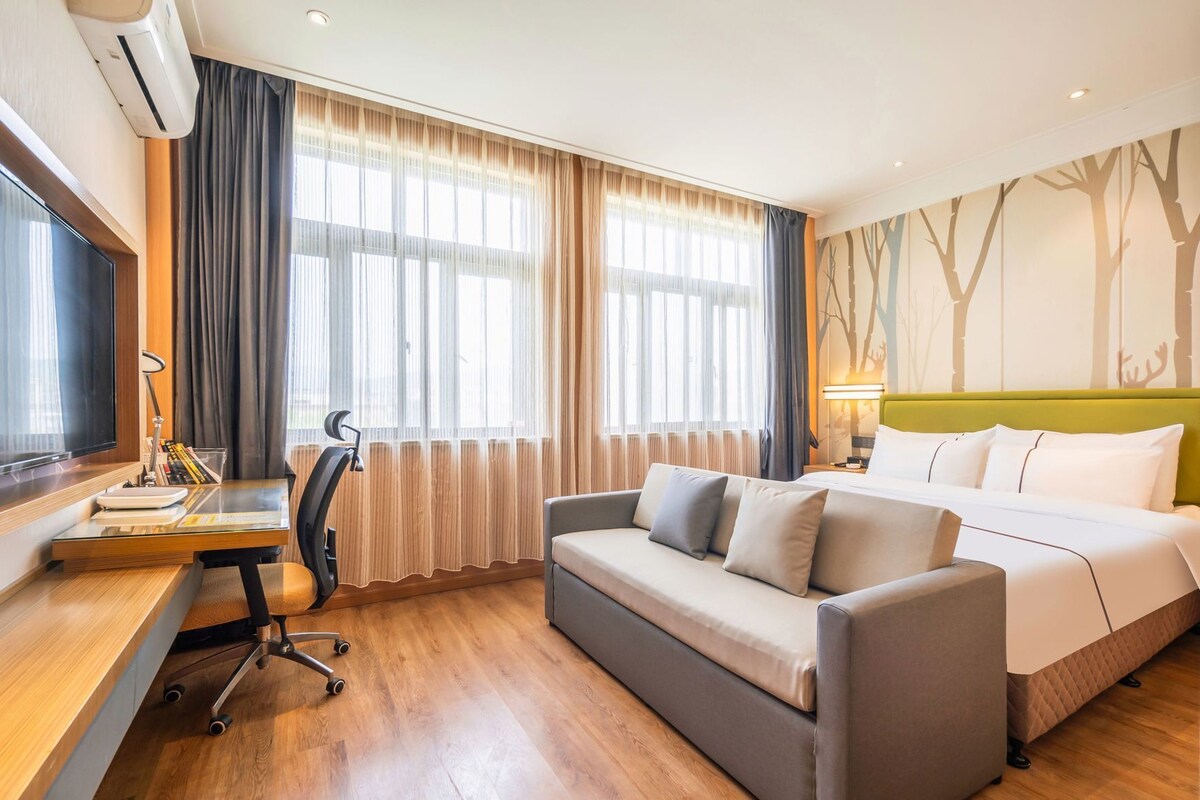 A hotel room with a bed, a sofa, a desk with a chair, a TV, and a window with curtains.