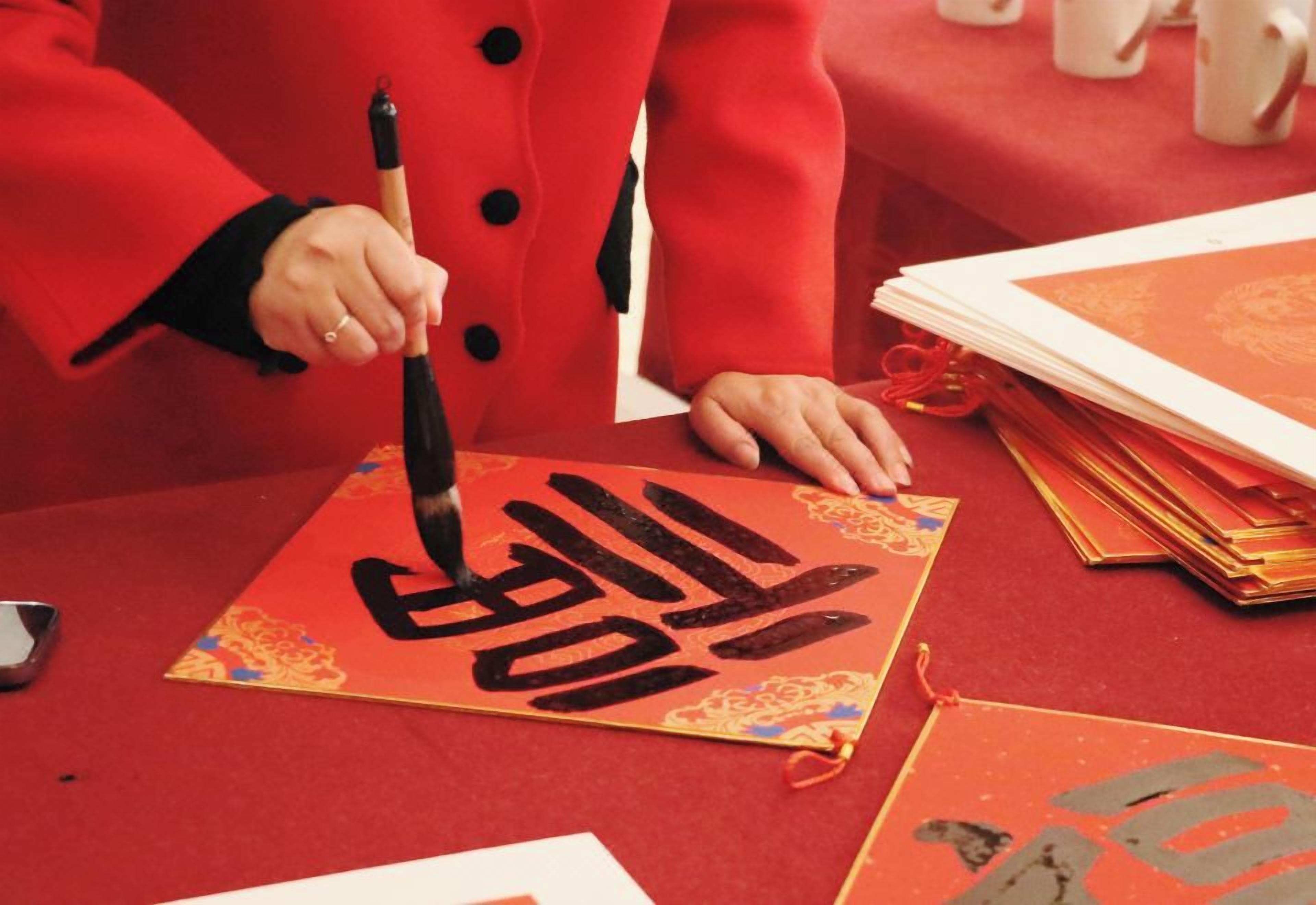 A person writing on a red envelope with a brush, surrounded by stacks of similar envelopes.