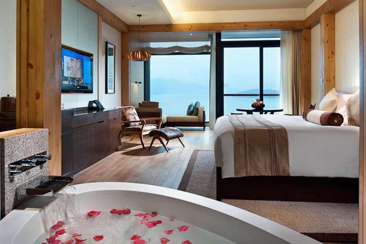 Habitación de hotel con una cama grande, una zona de estar con sillas, un escritorio, una bañera con pétalos de rosa y vistas a las montañas.