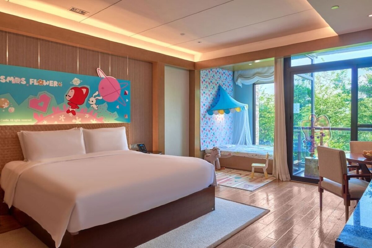 Habitación de hotel moderna con una cama grande, zona de comedor con mesa y sillas, ventanal con vistas a zonas verdes y decoración mural colorida.