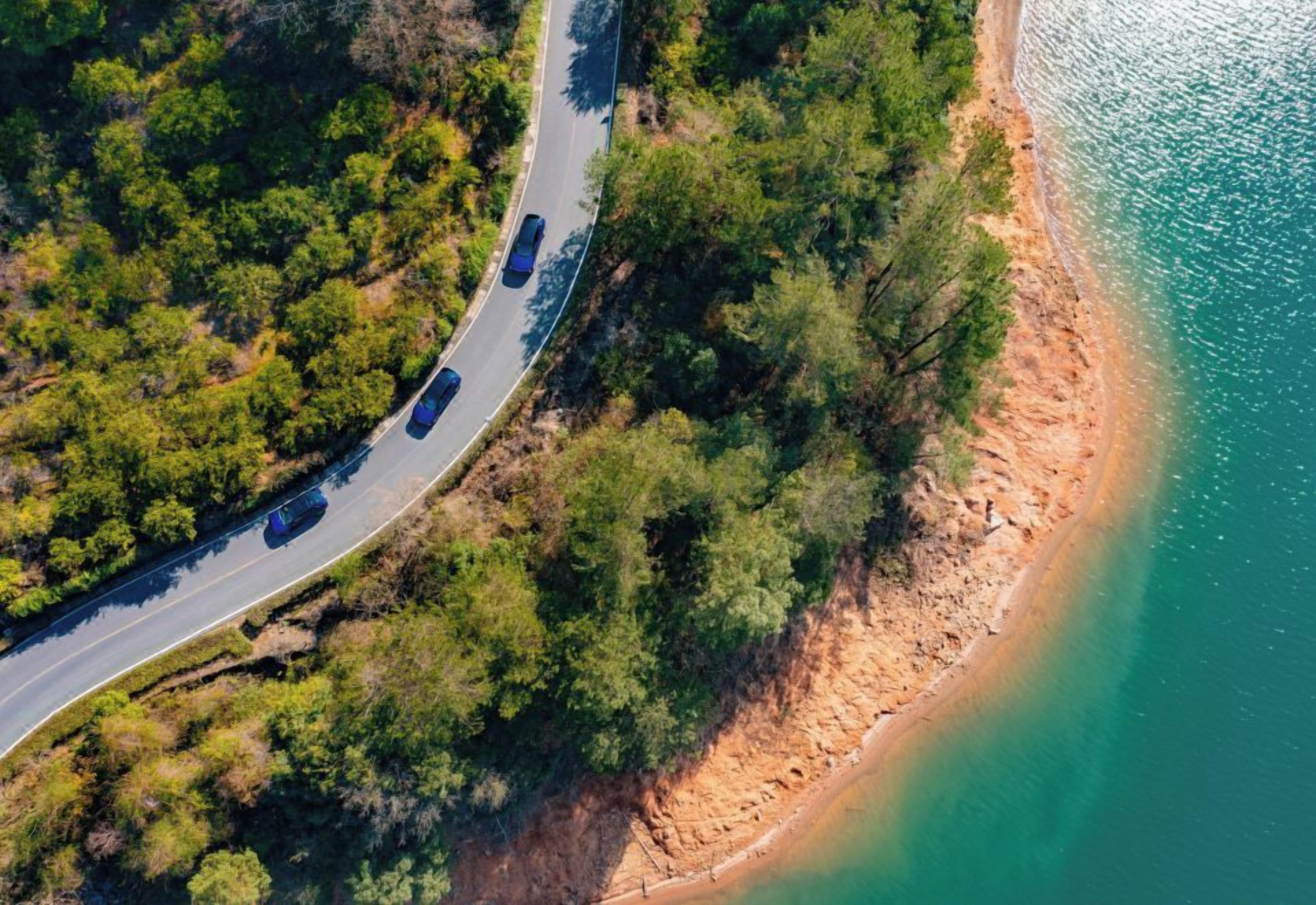 Vue aérienne d’une route côtière bordée de véhicules, entourée d’une végétation luxuriante et offrant une vue dégagée sur le littoral.