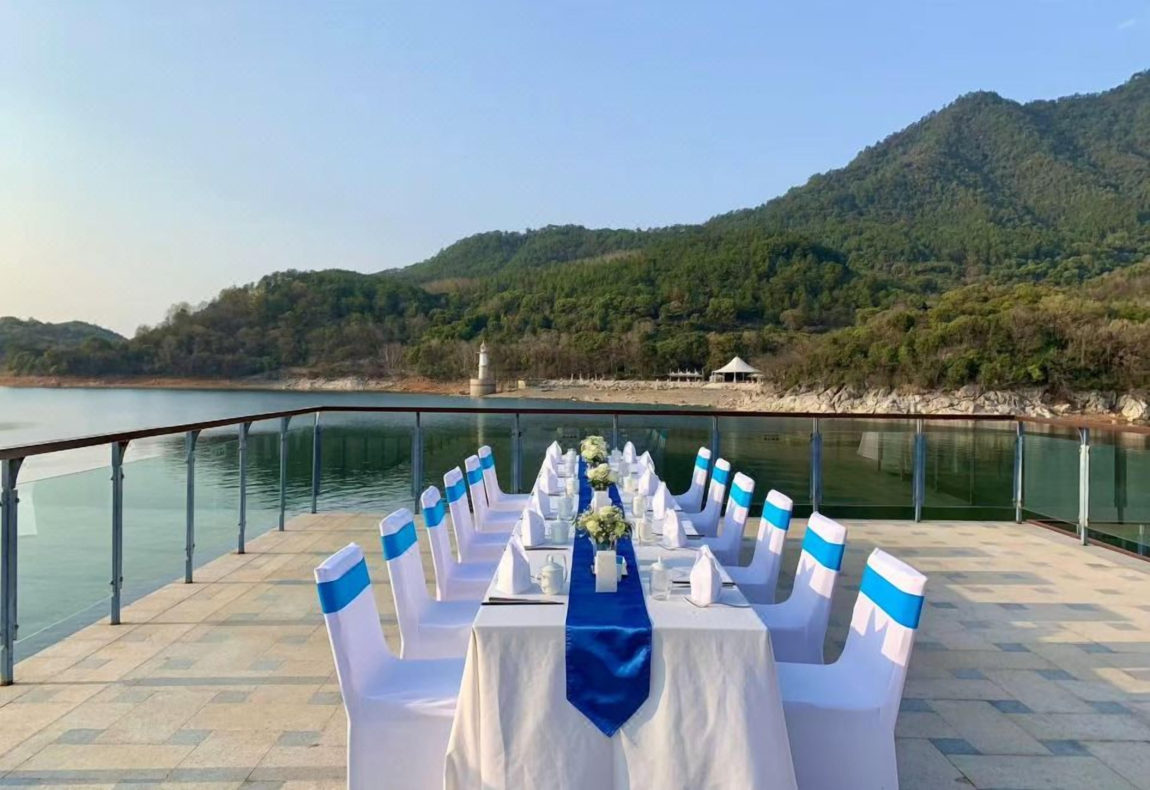 Une table est installée avec des chaises sur une terrasse au bord du lac, d’où l’on a une vue sur une colline boisée.