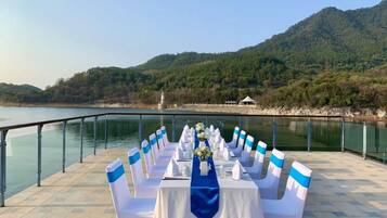 A table set up with chairs along a lakeside terrace, overlooking a forested hillside.