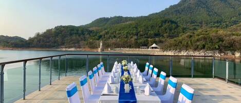 A table set up with chairs along a lakeside terrace, overlooking a forested hillside.