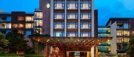 A modern hotel with a large glass facade, a covered outdoor area with a sculpture, and a pool.