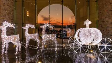 A festive display with illuminated reindeer and a sleigh, set against a backdrop of a sunset through a large window.