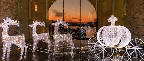 A festive display with illuminated reindeer and a sleigh, set against a backdrop of a sunset through a large window.