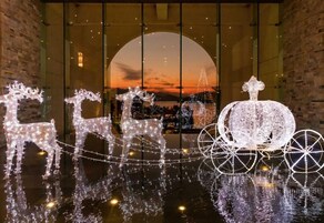 A festive display with illuminated reindeer and a sleigh, set against a backdrop of a sunset through a large window.