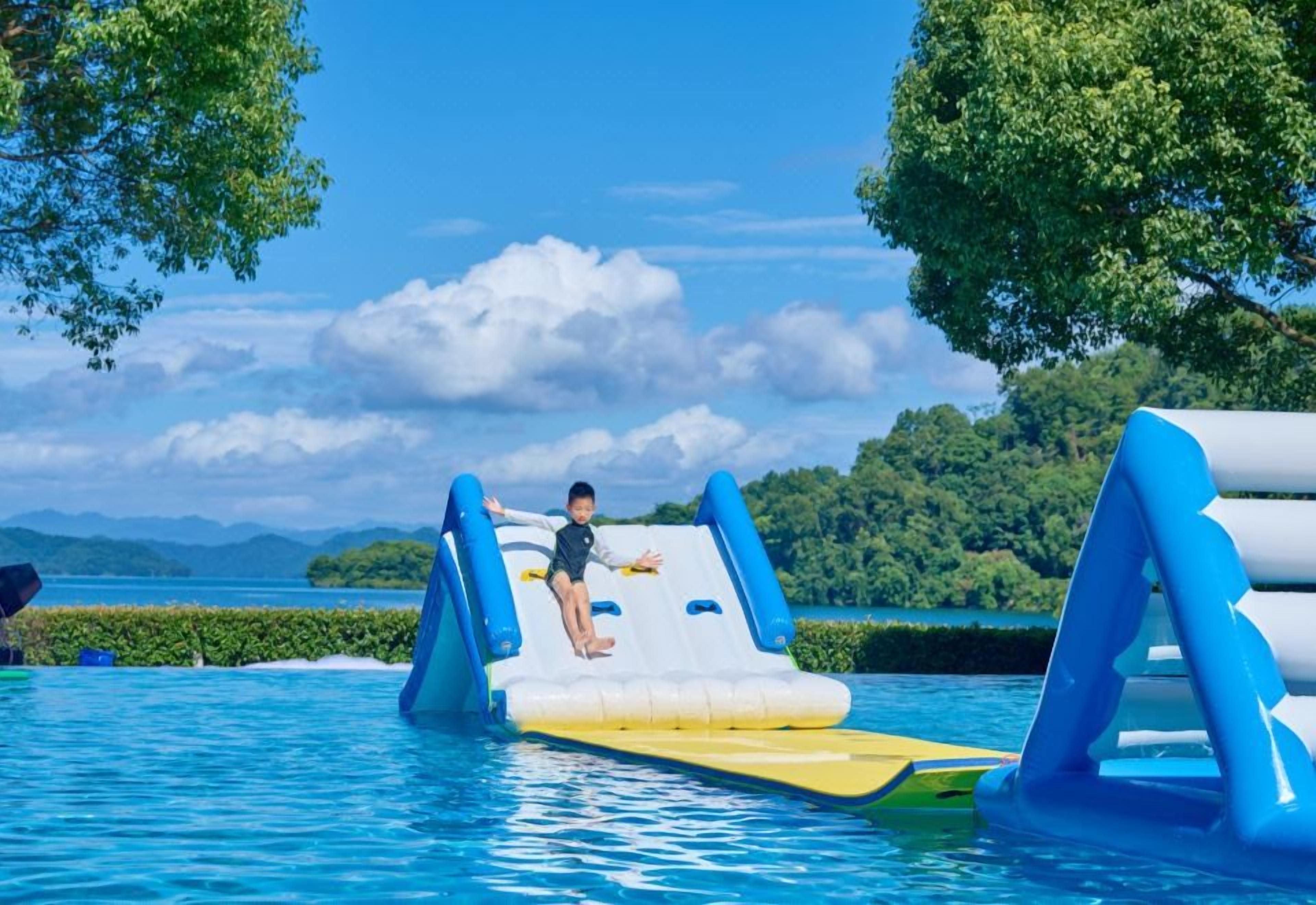 Ein Kind rutscht auf einer Wasserrutsche in einem Pool, im Hintergrund ein klarer Himmel und Berge.