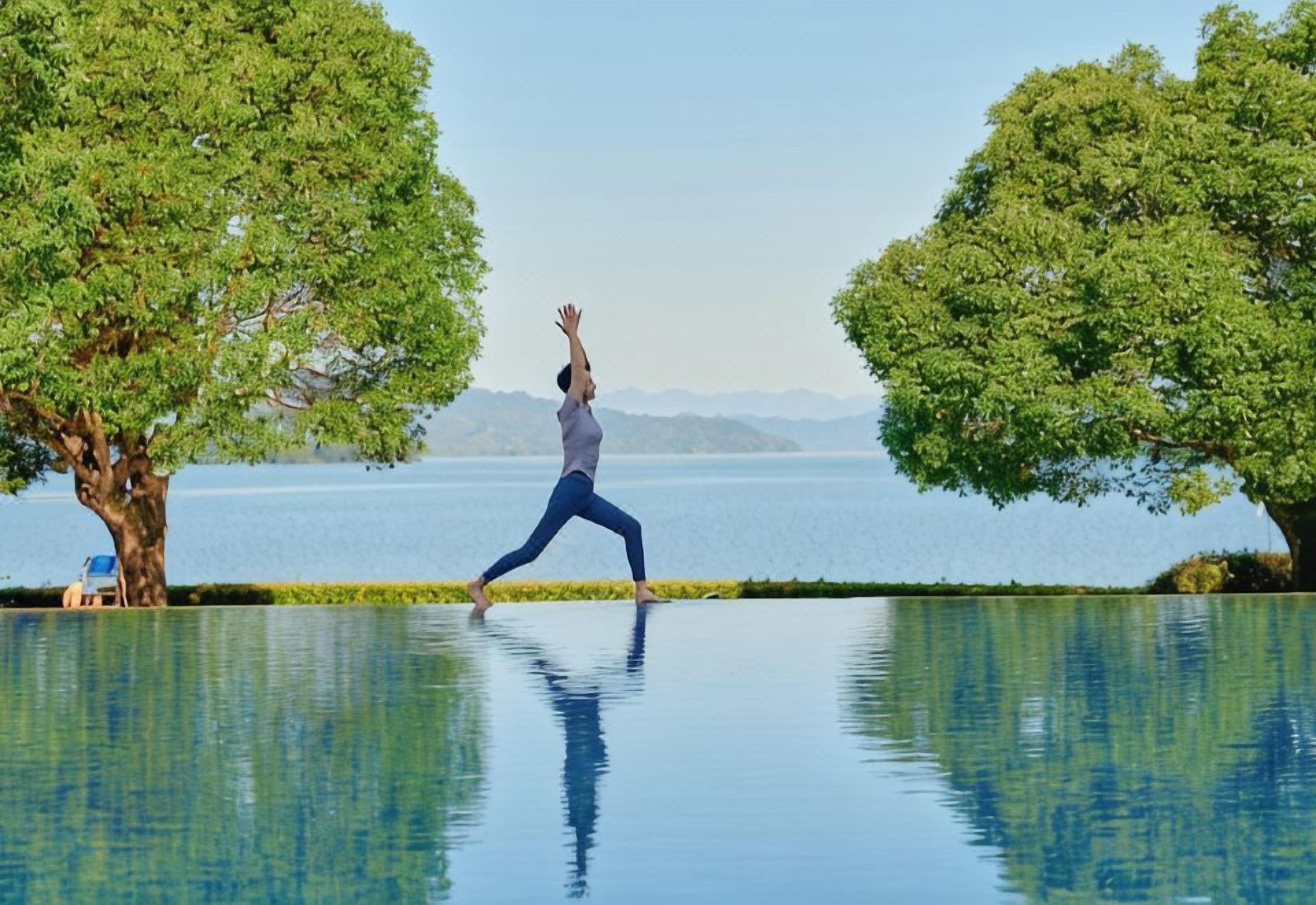A person performing a yoga pose on a reflective pool with trees and a distant view of mountains.