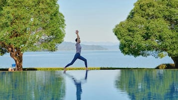 A person performing a yoga pose on a reflective pool with trees and a distant view of mountains.