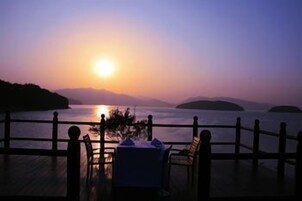 Vista del atardecer desde una terraza con mesa y sillas, con vista a una masa de agua tranquila y formaciones terrestres a lo lejos.