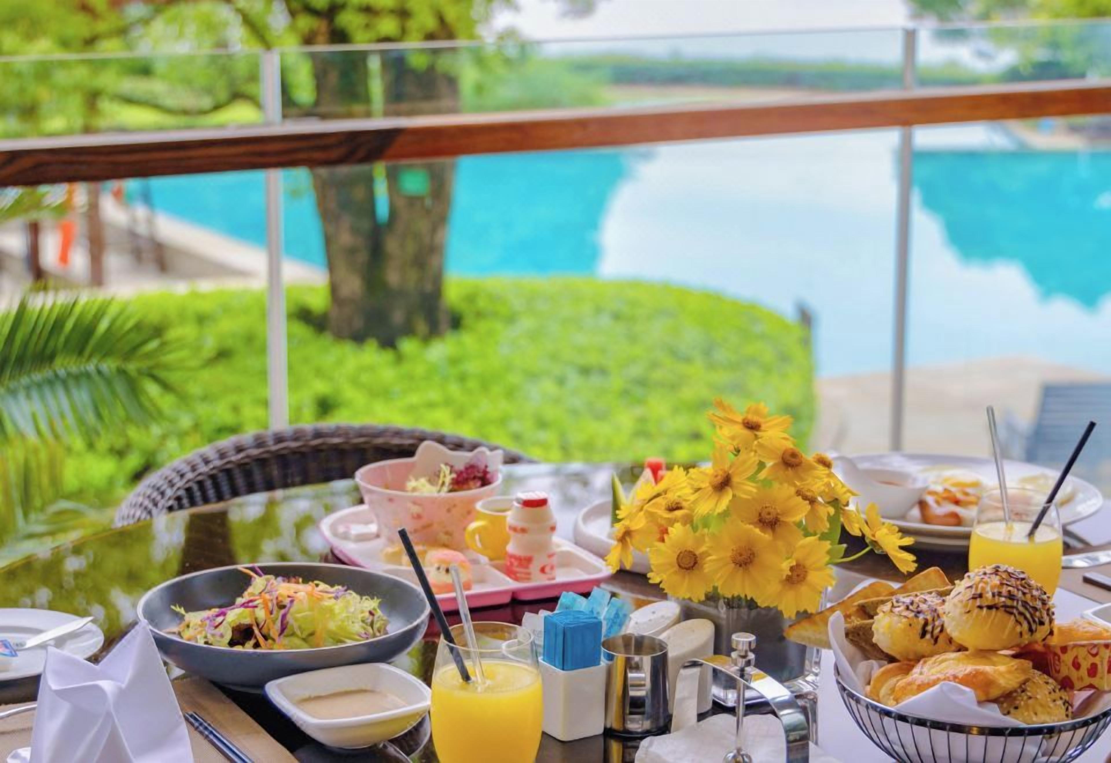 Une table de petit-déjeuner avec différents plats, un vase rempli de fleurs jaunes et une vue sur une étendue d’eau.