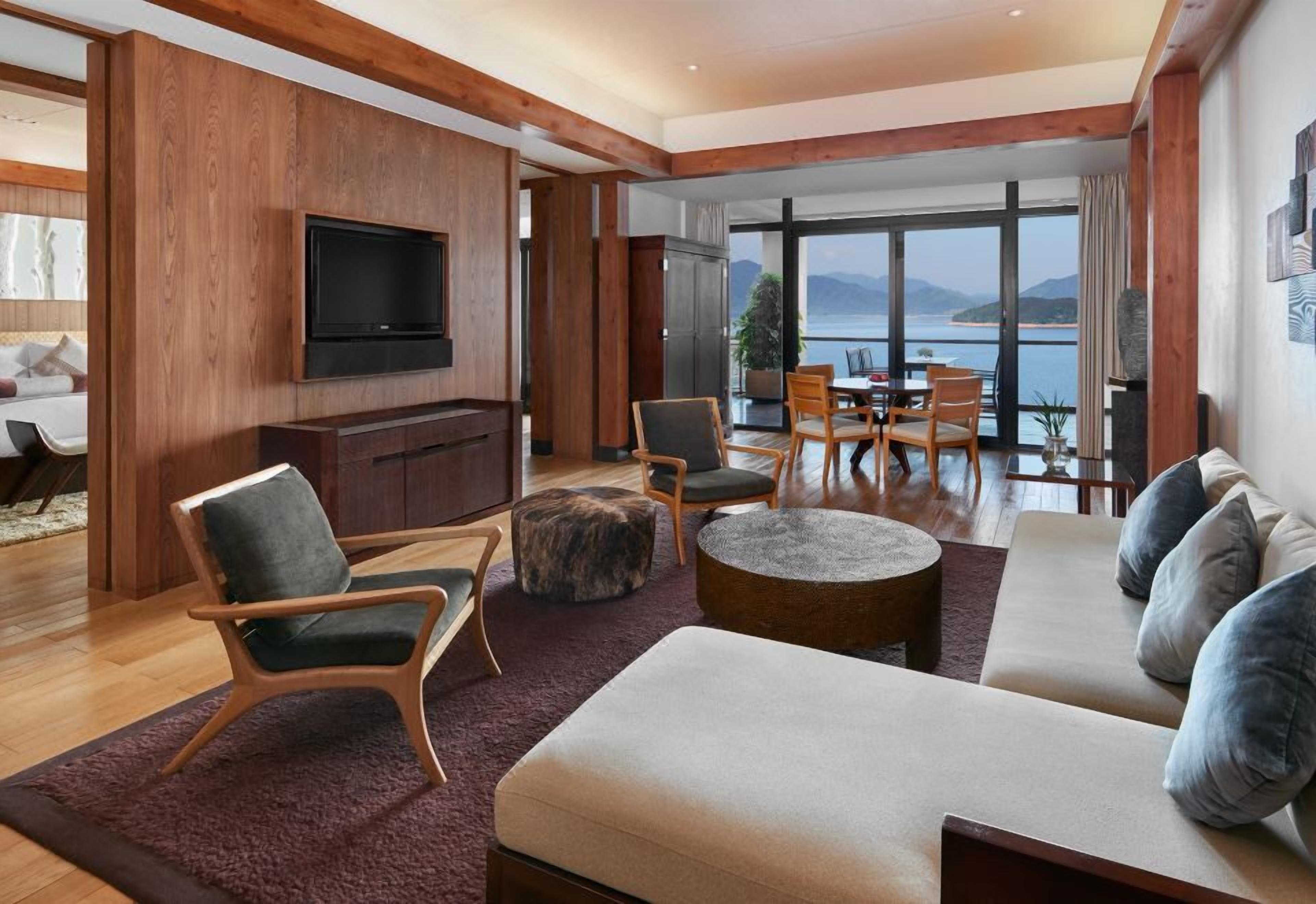 A modern living room with a large TV, comfortable seating, a dining area, and a view of the sea.
